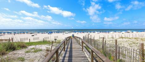 A wooden boardwalk leading to a sandy beach with umbrellas and beachgoers.