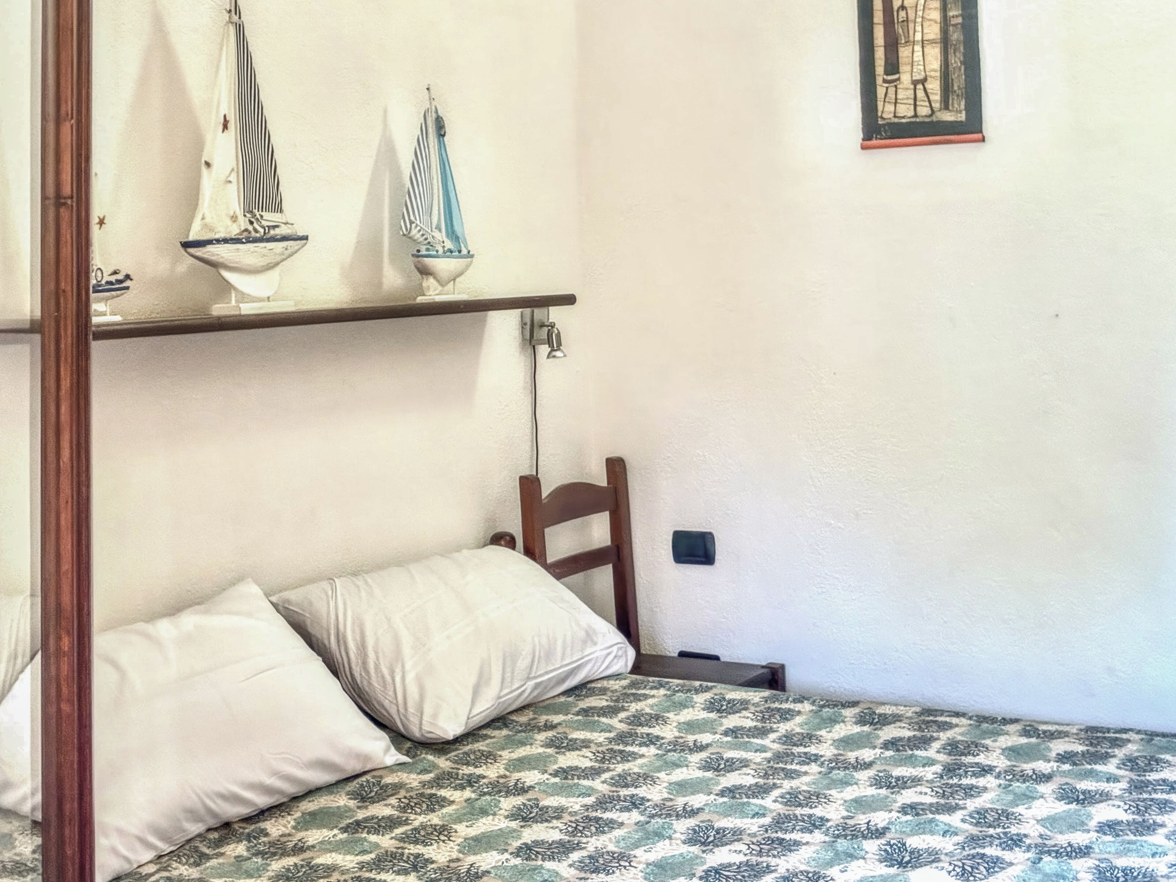 A bed with a patterned bedspread, two pillows, a wooden chair, and two wall-mounted shelves with sailboat decorations.