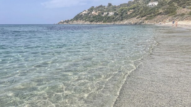 En strand med klart vatten, en sandstrand och hus som ligger på en sluttning.