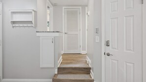 A hallway with a wooden staircase, white walls, a white door with a louvered design, and a small white shelf. - Near Beach Coastal Getaway (Hilton Head Island)