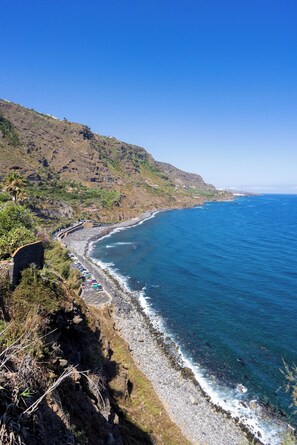 A coastal road with a rocky shoreline, a clear blue sky, and a hilly landscape.