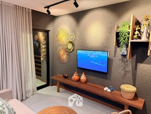 A modern living room with a flat-screen TV mounted on the wall, a wooden bench with decorative items, and a large window with curtains.