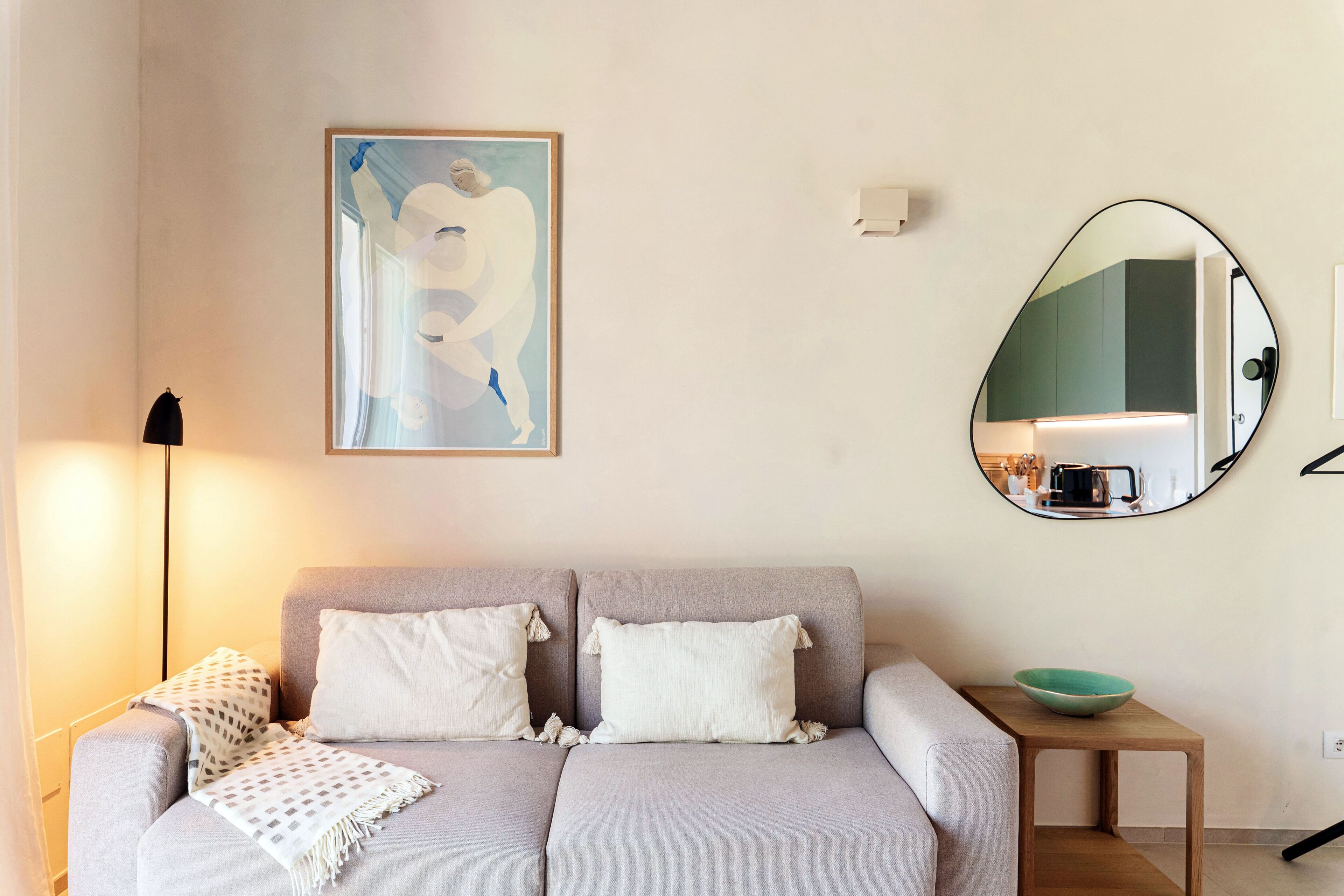 A living room with a sofa, pillows, a throw blanket, a small table with a bowl, a framed abstract painting, and a mirror reflecting a kitchen area.