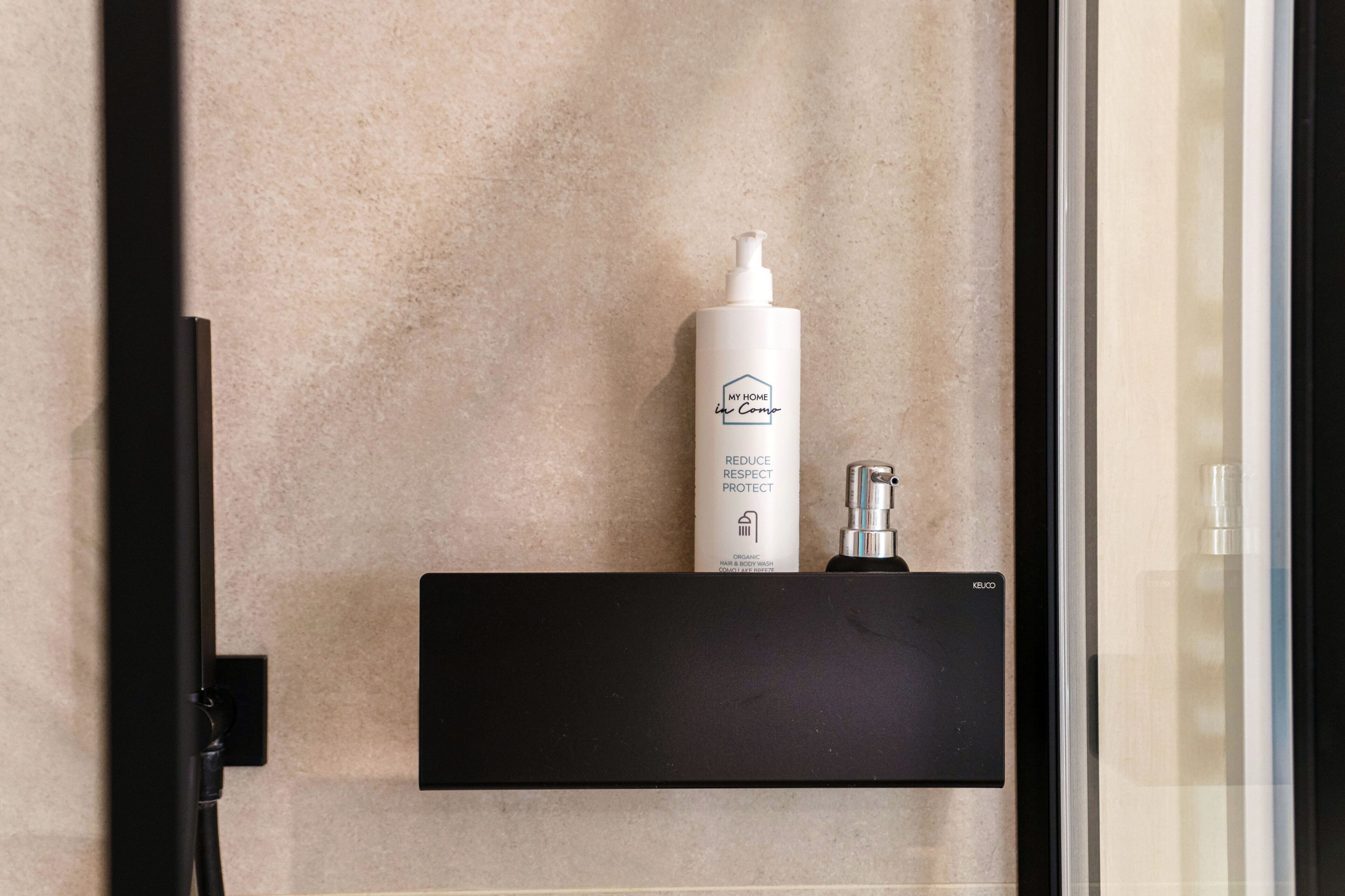A bathroom with a beige wall, a black shelf holding a white bottle labeled 'My Home' and a black object.