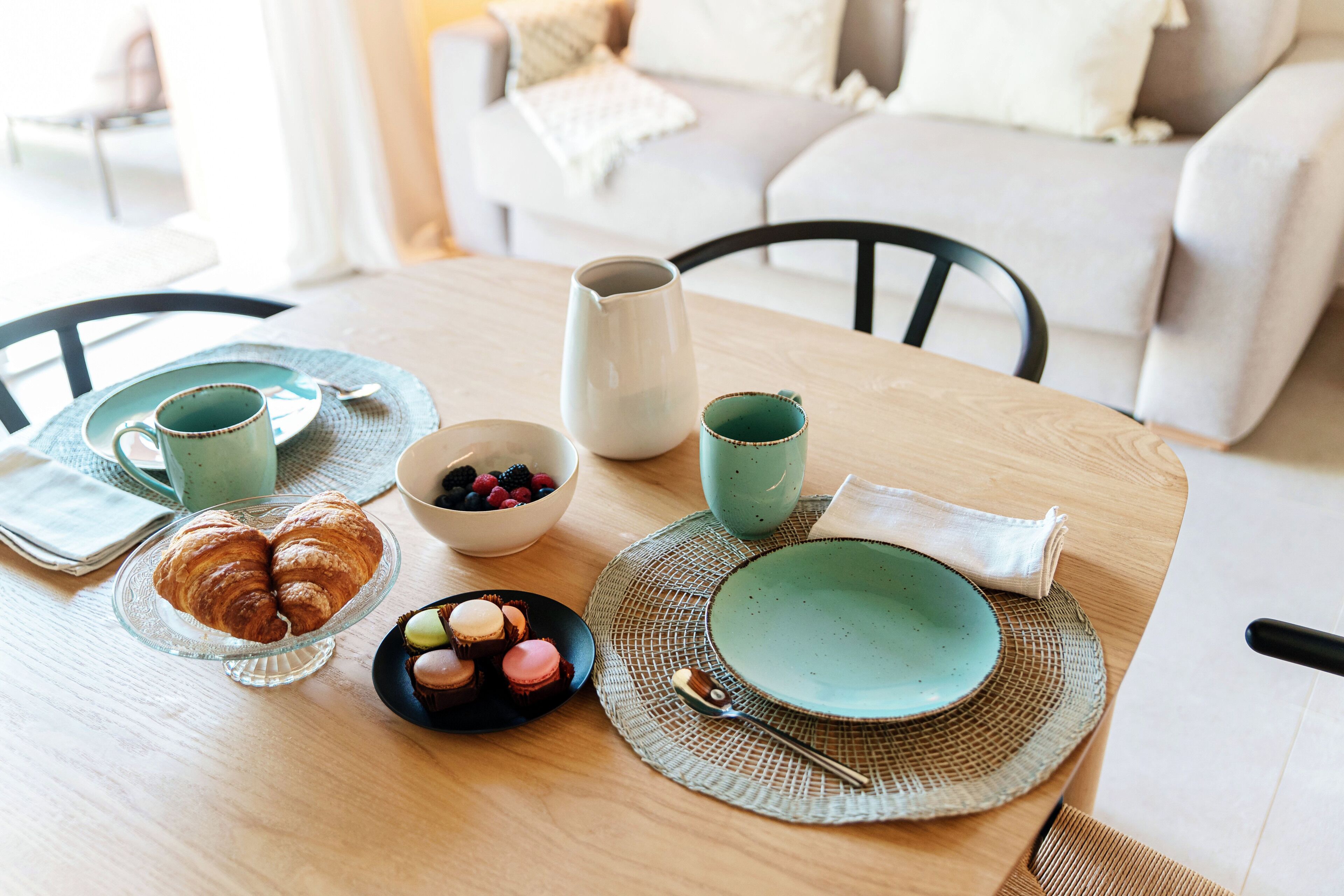 A dining table with a bowl of fruit, croissants, and macarons, set with green plates and a pitcher.