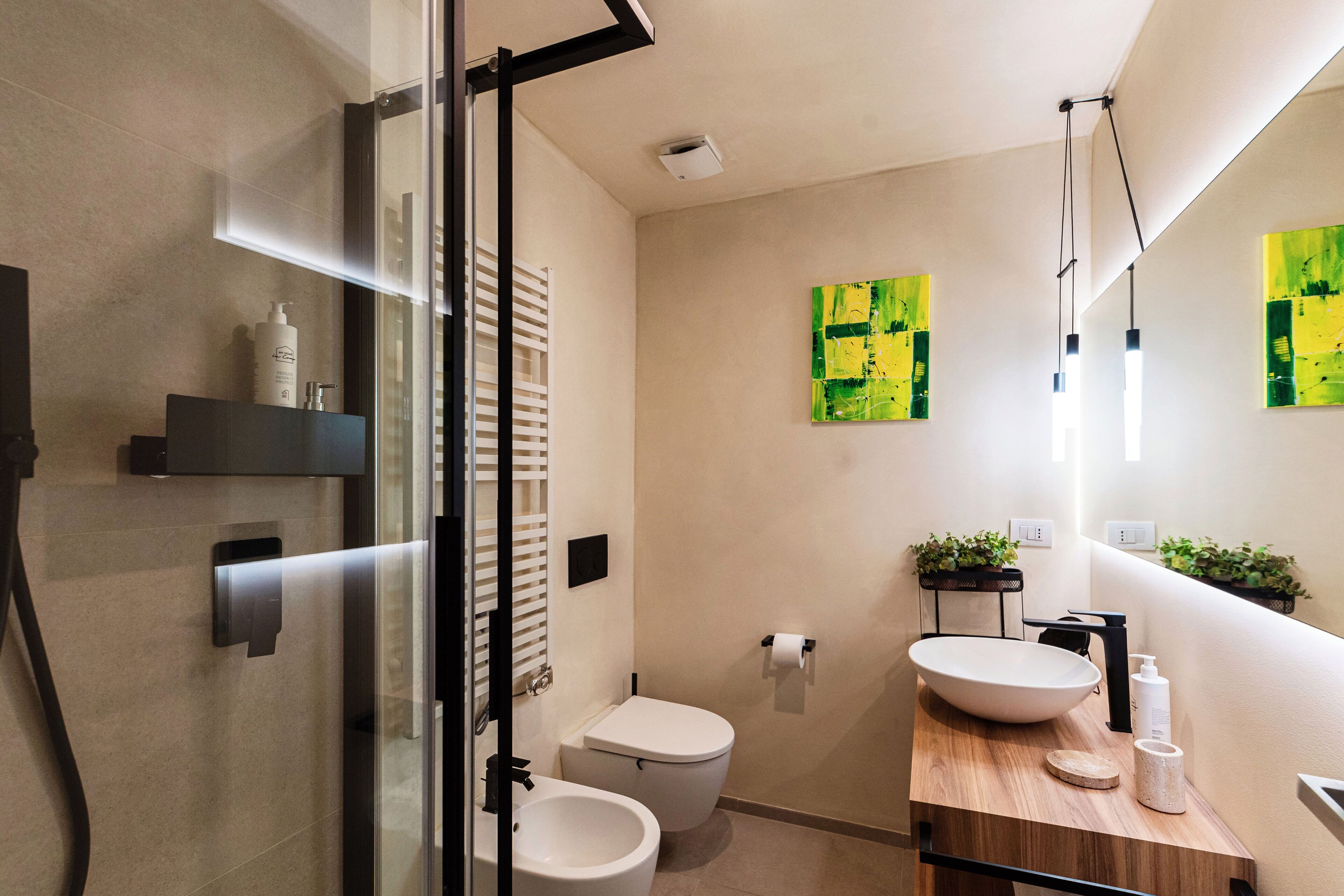 A modern bathroom with a glass shower enclosure, a toilet, a sink with a wooden countertop, and two green abstract paintings on the wall.