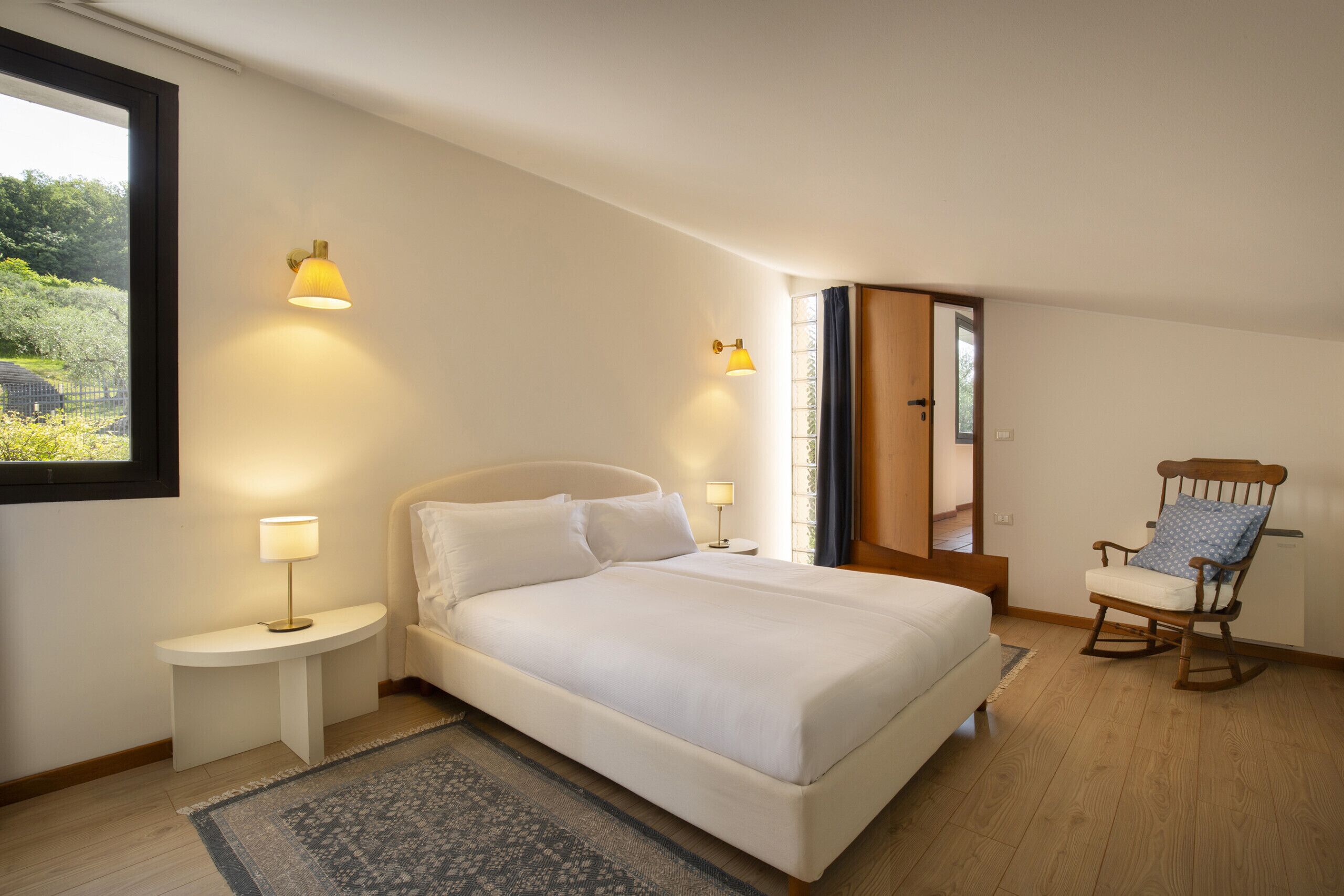 Un dormitorio con cama, mesitas de noche, una mecedora y vistas al exterior.