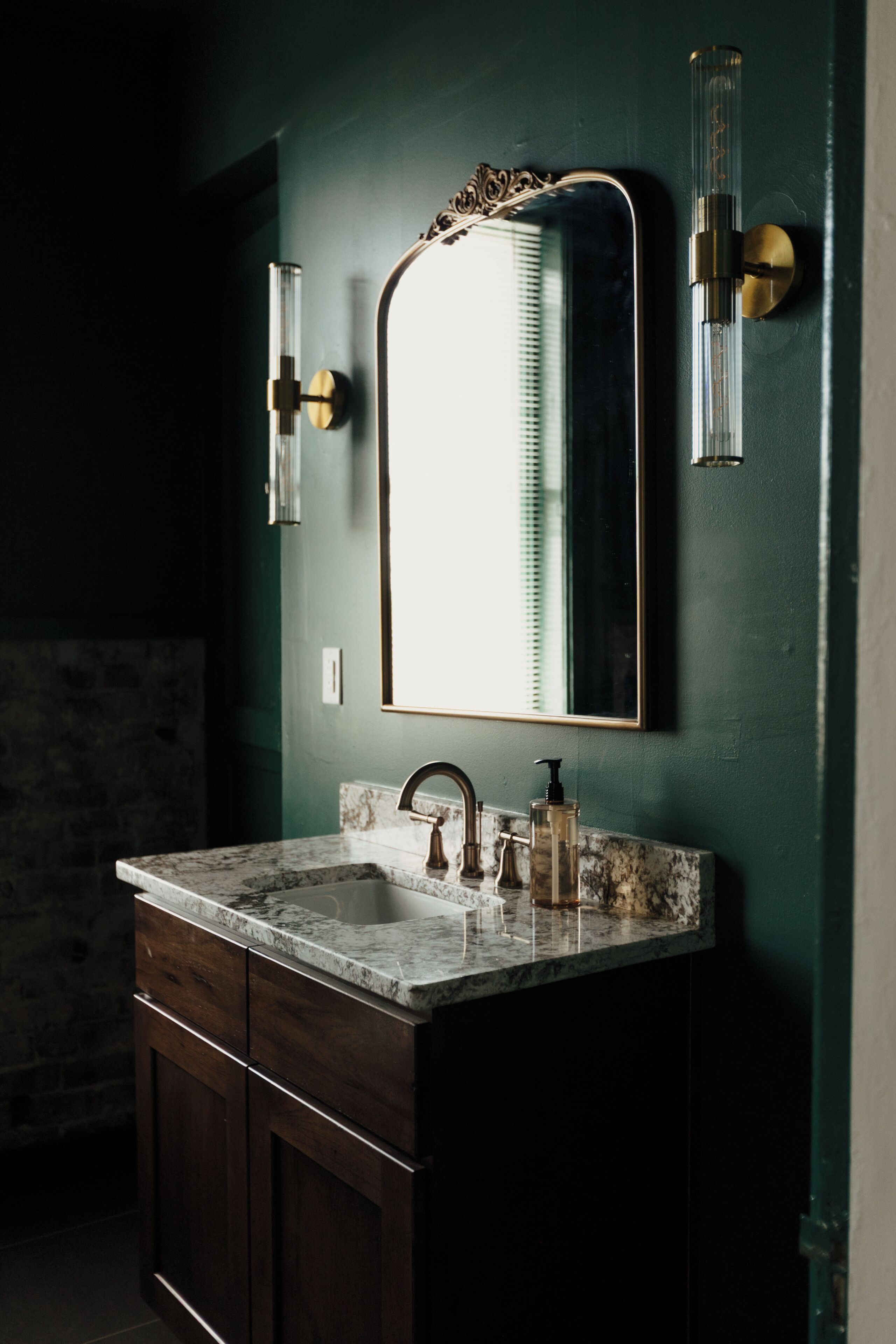 A bathroom with a marble sink, dark wooden cabinets, a large mirror, and a wall-mounted light fixture.