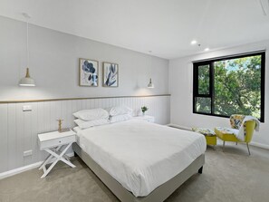 A bedroom with a bed, bedside tables, a chair, a window with a view of trees, and wall art. - Scenic Batemans Bay Stay Near Golf Course (Batemans Bay)