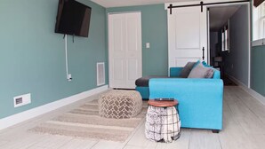 A living room with teal walls, a blue sofa, a patterned ottoman, a small round table, and a TV mounted on the wall. - Oak Island Sun: 3 blocks to beach, fenced yard, fire pit (Oak Island)