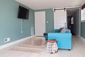 Ein Wohnzimmer mit türkisfarbenen Wänden, einem blauen Sofa, einem gemusterten Hocker, einem kleinen runden Tisch und einem an der Wand montierten Fernseher.
