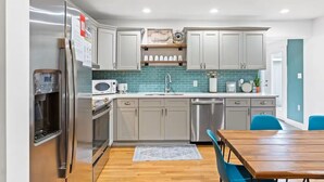 A modern kitchen with stainless steel appliances, grey cabinets, a teal backsplash, and a wooden dining table. - Oak Island Sun: 3 blocks to beach, fenced yard, fire pit (Oak Island)