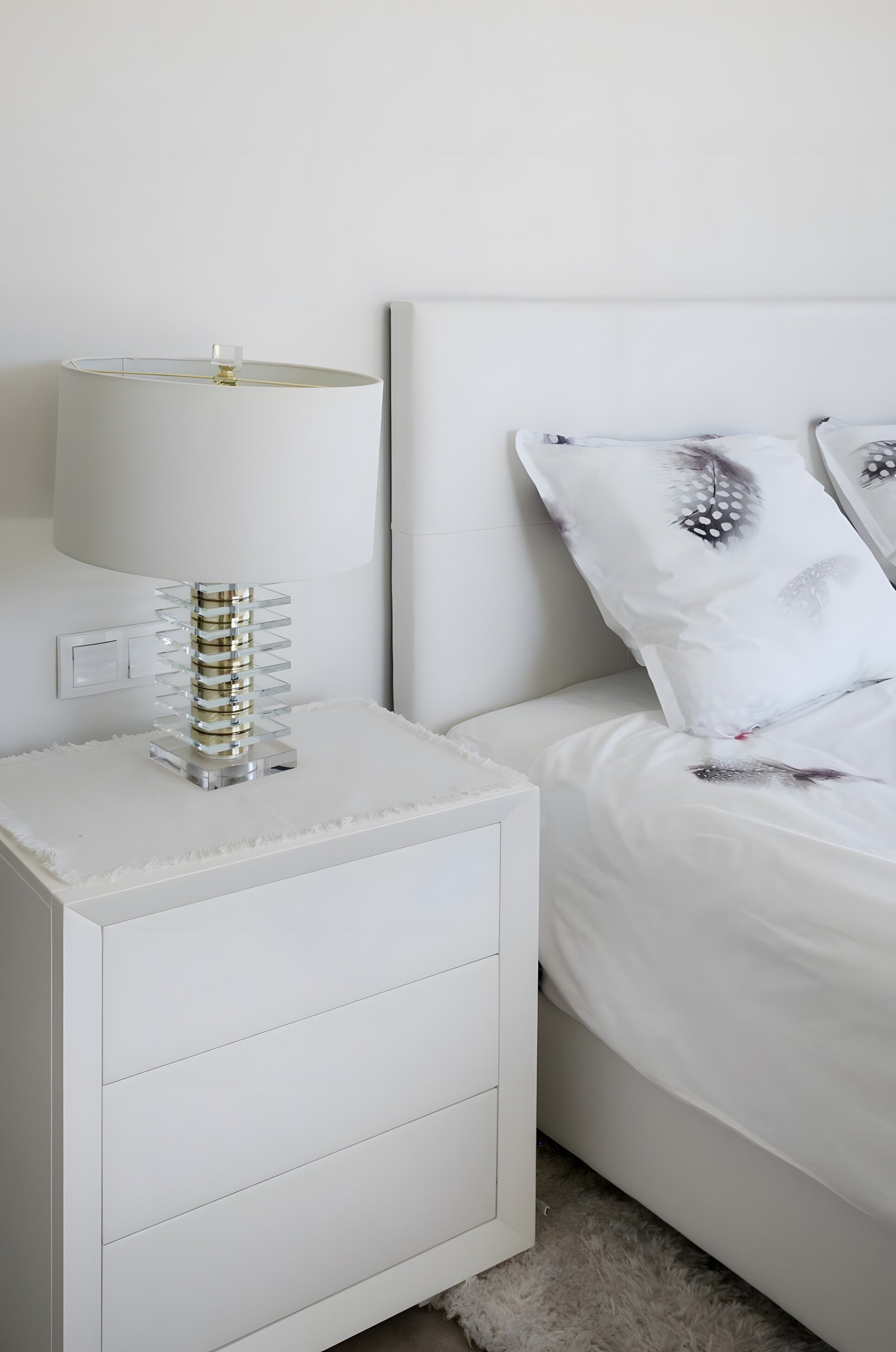 A white bedside table with a lamp and a decorative glass structure, a white bed with patterned pillows, and a white nightstand with drawers.