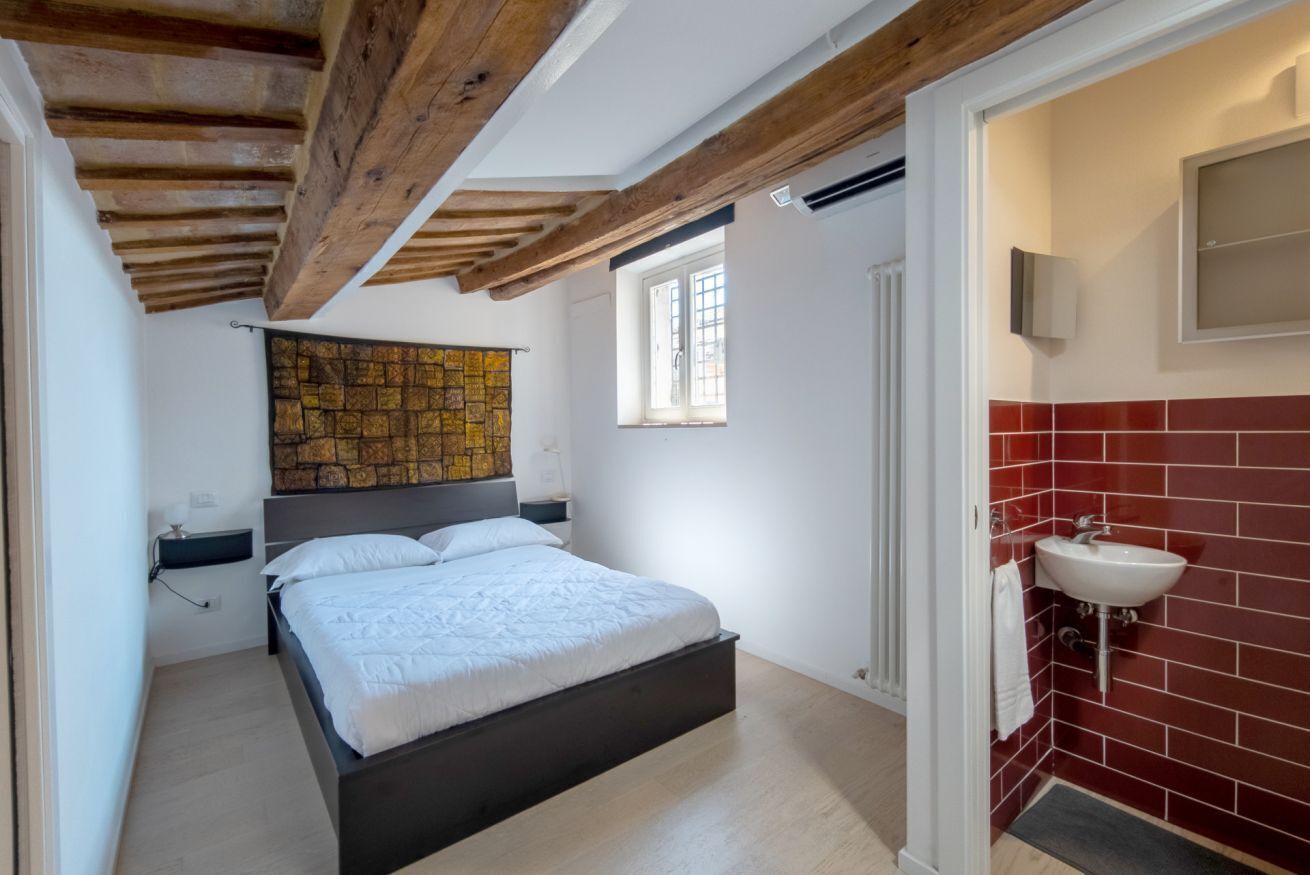 A bedroom with a bed, a wall-mounted artwork, a window, and a bathroom with a sink and red-tiled walls.