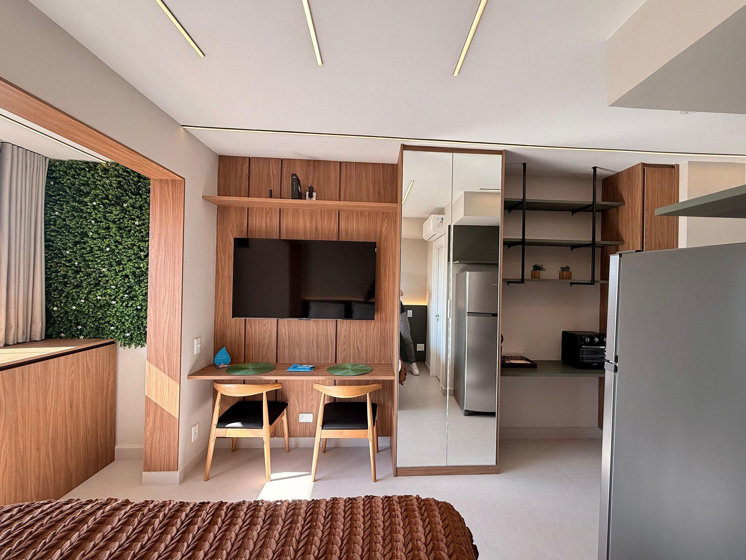 Una habitación moderna con cama, televisión, escritorio con sillas, zona de cocina y pared verde.