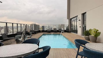 En pool på taket med solstolar och bord, utsikt över staden och en byggnad med stora fönster.