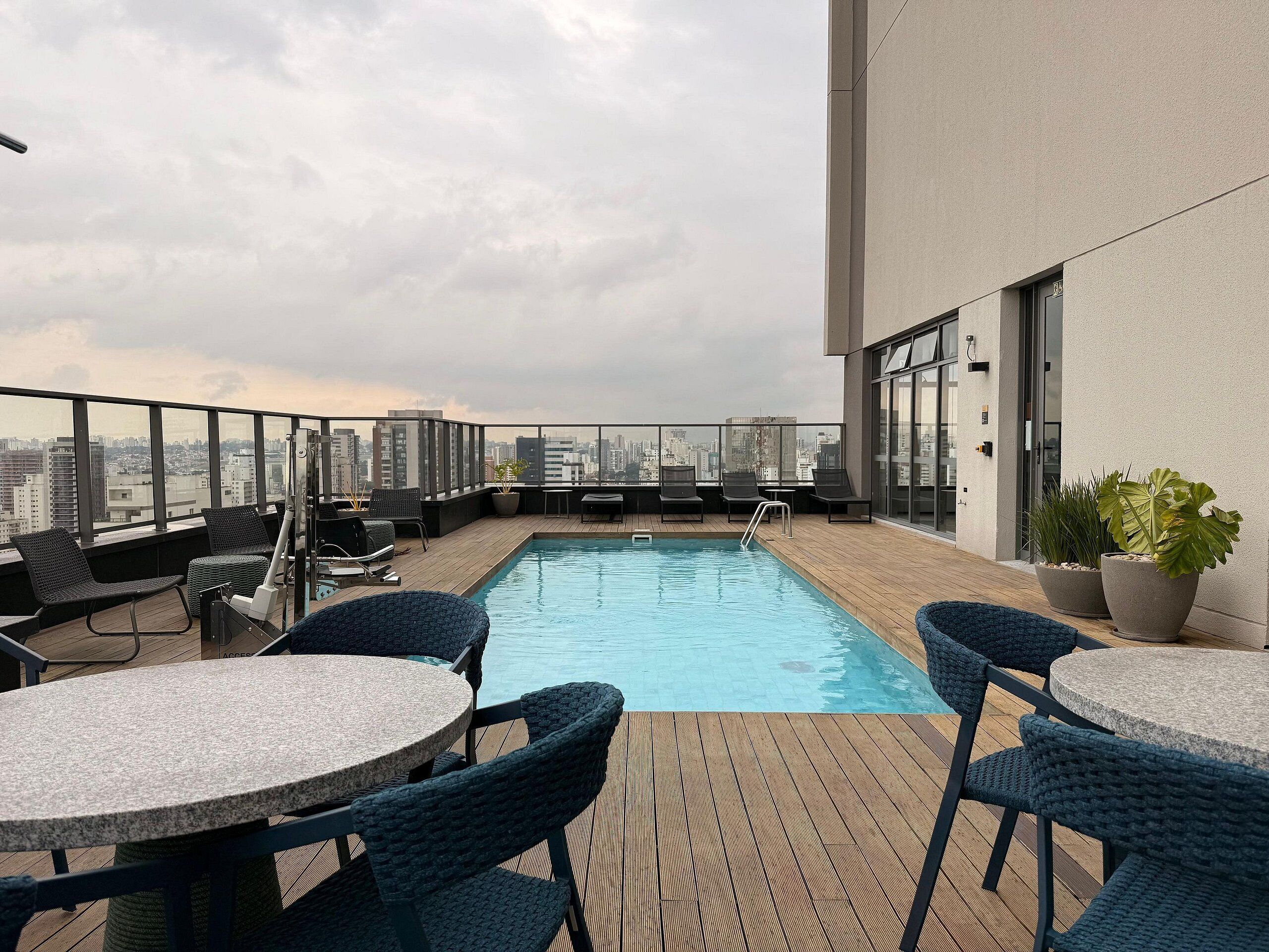 A rooftop pool with lounge chairs and tables, a city view, and a building with large windows.