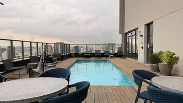 A rooftop pool with lounge chairs and tables, a city view, and a building with large windows.