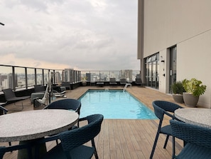 Una piscina en la azotea con sillones y mesas, vistas a la ciudad y un edificio con amplias ventanas.