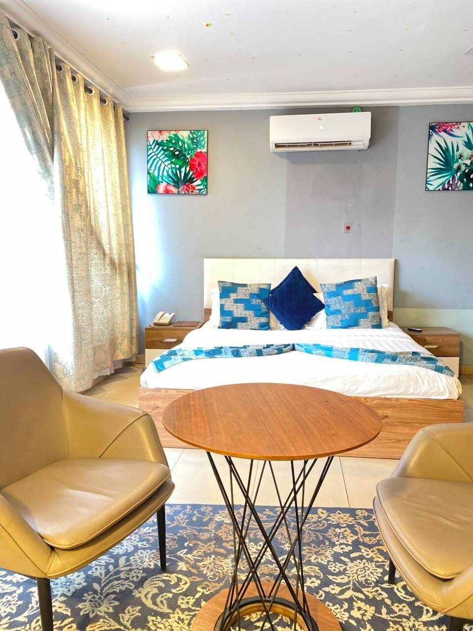 A hotel room with a bed, two chairs, a small table, an air conditioning unit, and decorative wall art.