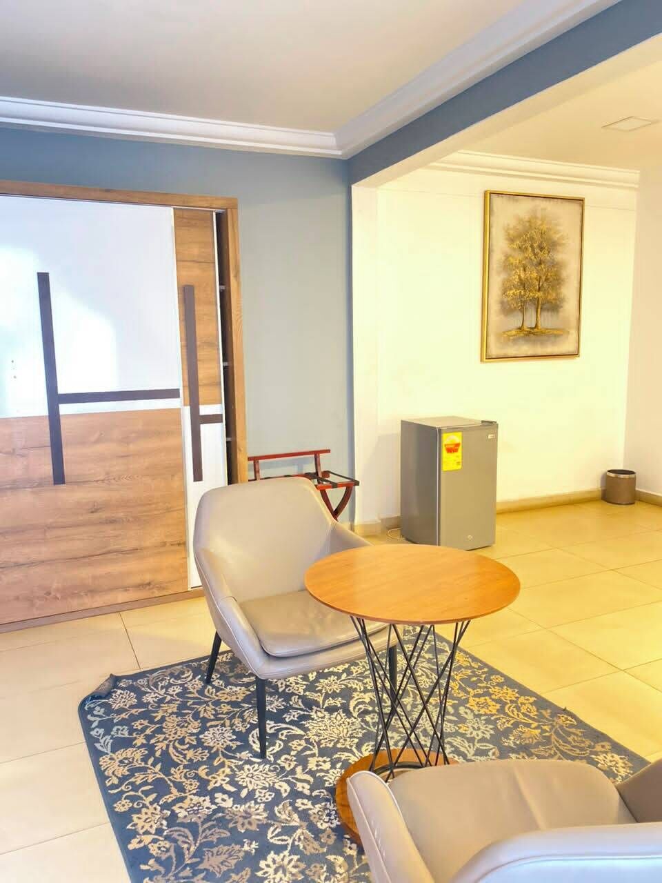 A room with a chair, a small round table, a patterned rug, a refrigerator, and a framed picture on the wall.