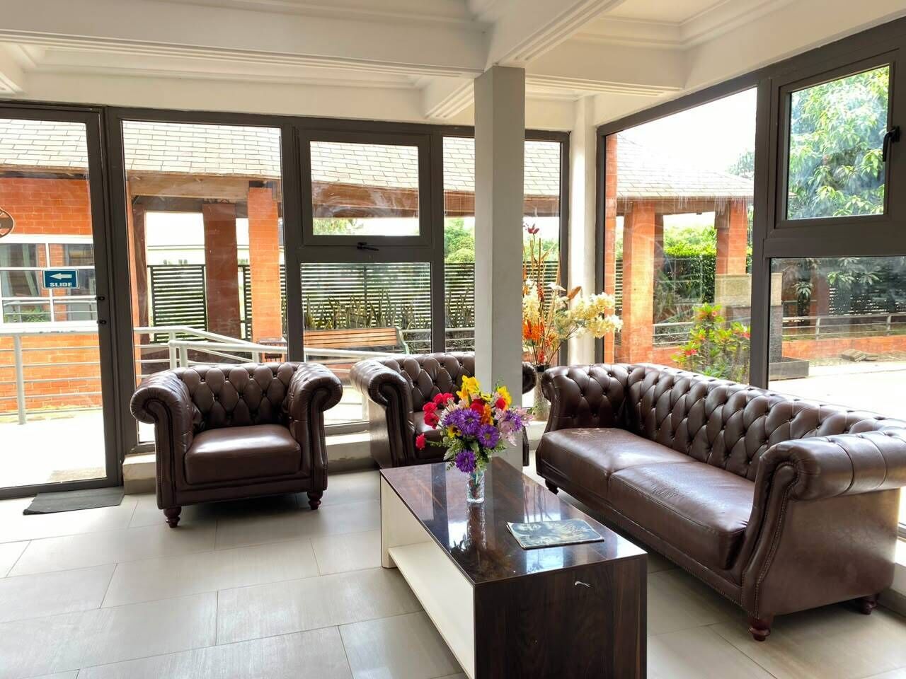 A spacious room with a brown leather sofa, two armchairs, and a coffee table with a vase of flowers. Large windows provide a view of the outdoors.