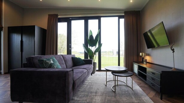 A modern living room with a sofa, a coffee table, a TV, and a large window with curtains. - Holiday Home in Kampen With Terrace (Kampen)