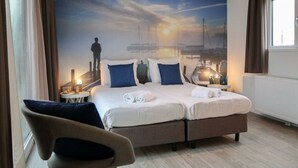 Habitació d'hotel amb un llit, una cadira, una taula de nit i un mural d'un capvespre sobre un port.