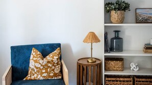 Et værelse med en blå polstret stol, et lille træsidebord med en lampe og en hvid reol med diverse dekorative genstande.