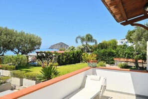 Ein Balkon mit Meer- und Bergblick, umgeben von Grünflächen und einem weißen Gebäude.