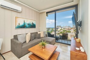 Un salon moderne avec un canapé gris, une table basse agrémentée d’une plante, un tableau au mur et une vue sur la ville et l’océan, visible à travers une grande porte-fenêtre coulissante.