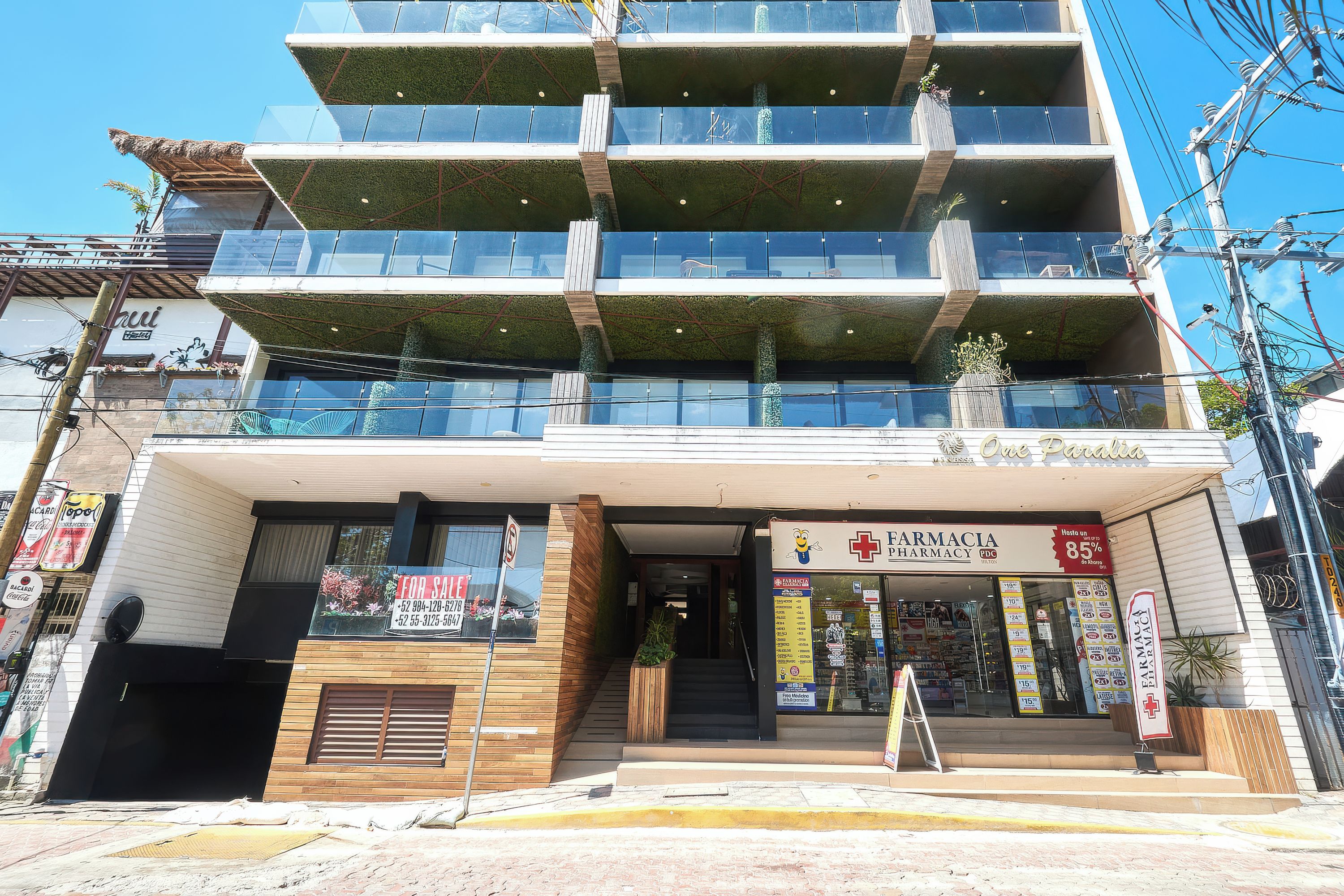A multi-story building with a pharmacy on the ground floor, a for sale sign, and a balcony with plants.