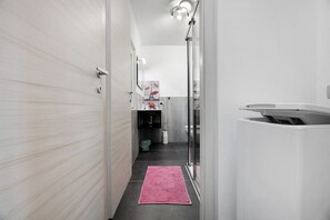 A narrow hallway with a pink rug, a glass shower enclosure, and a view of a bathroom with a toilet and sink.