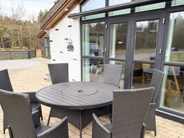 Un patio exterior con una mesa redonda y sillas de mimbre, una entrada con puerta de vidrio y una terraza de madera.