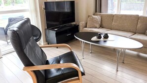 A living room with a black leather chair, a black entertainment unit with a TV, a beige sofa, and a round coffee table. - 9 Person Holiday Home in Saeby (Sæby)