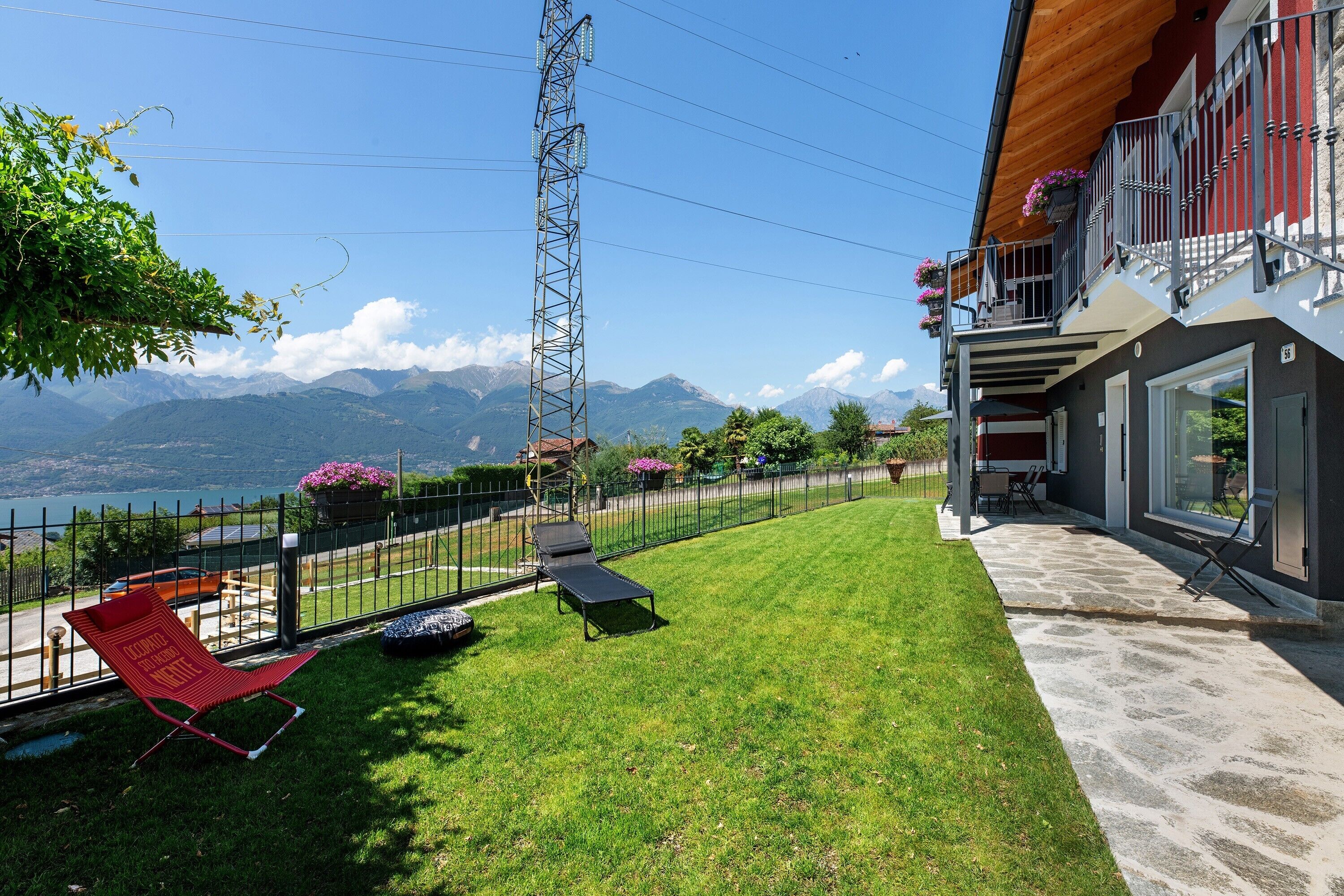 Una proprietà residenziale con vista su montagne e un lago, un giardino recintato con prato, una poltrona da relax rossa e un balcone con fioriere.