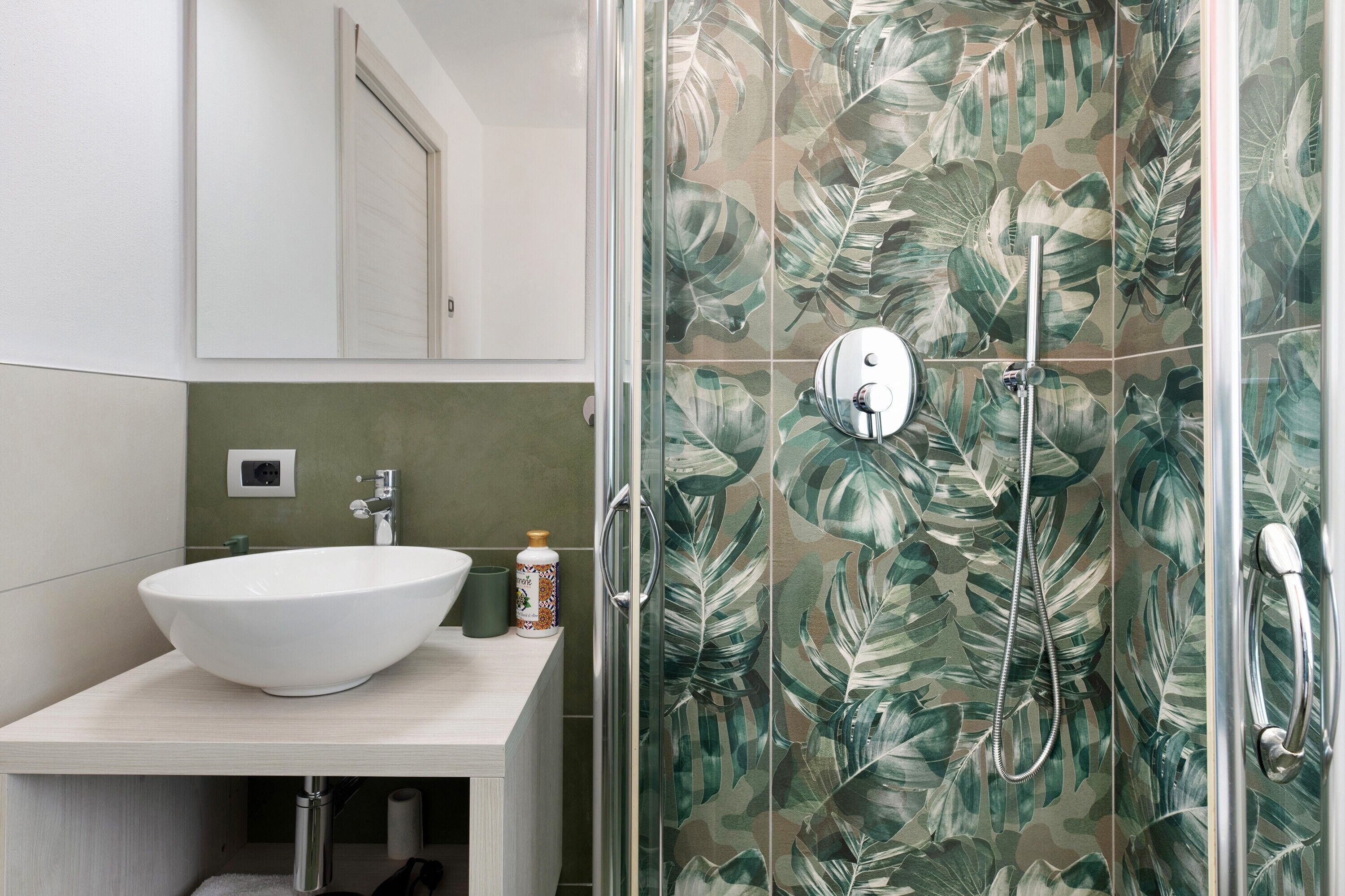 Un bagno con un lavandino bianco, una doccia con porta in vetro e una parete rivestita di piastrelle verdi.