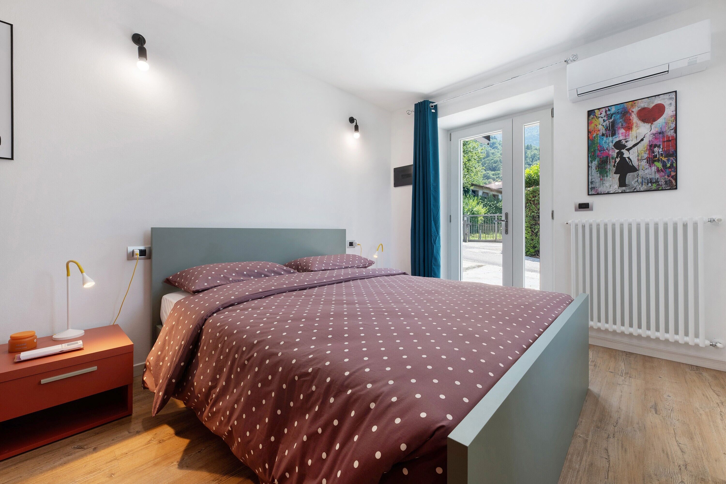 Un dormitorio con cama, mesita de noche, lámpara y vistas al exterior.