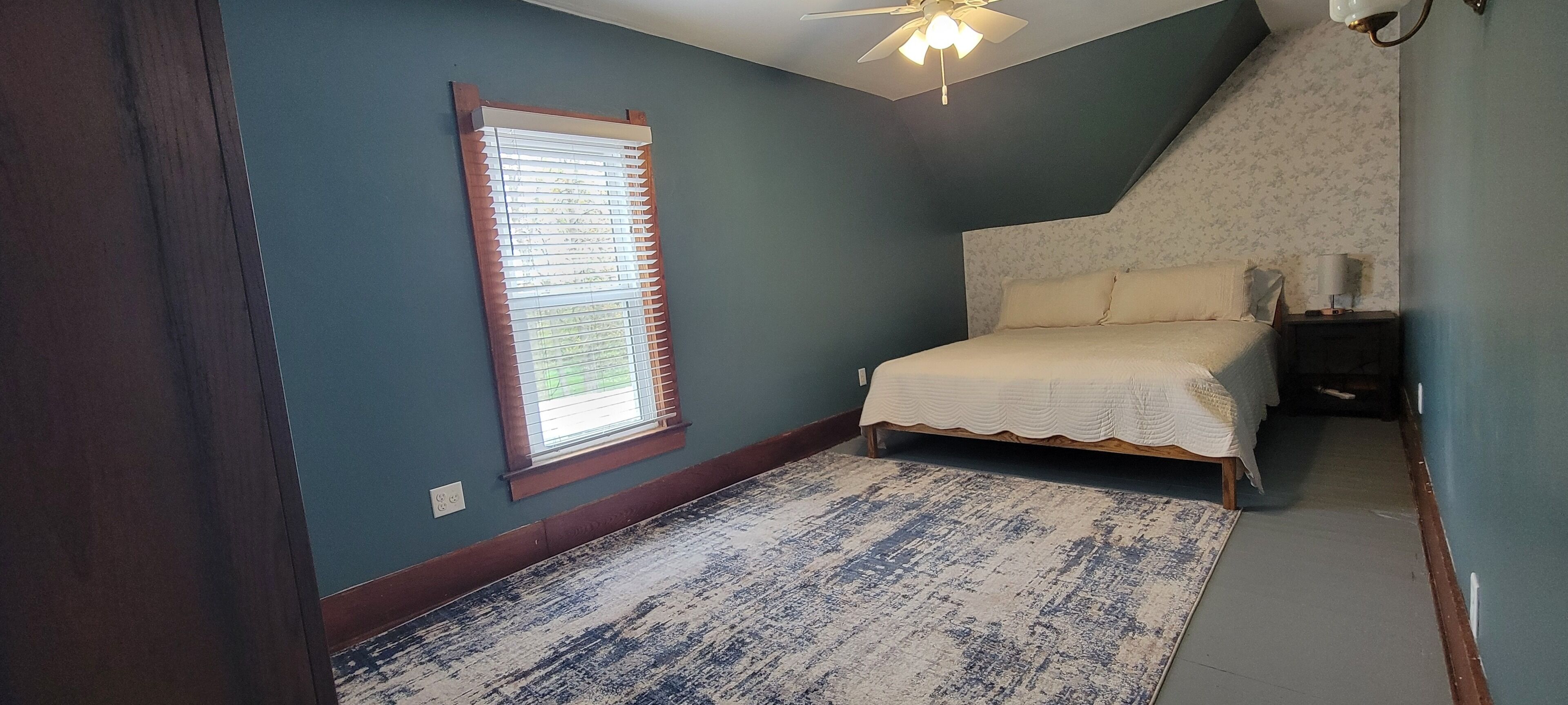 A bedroom with a bed, a window with blinds, a ceiling fan, and a patterned rug.