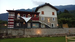 A two-story house with a balcony, stone wall, and a mountain backdrop.