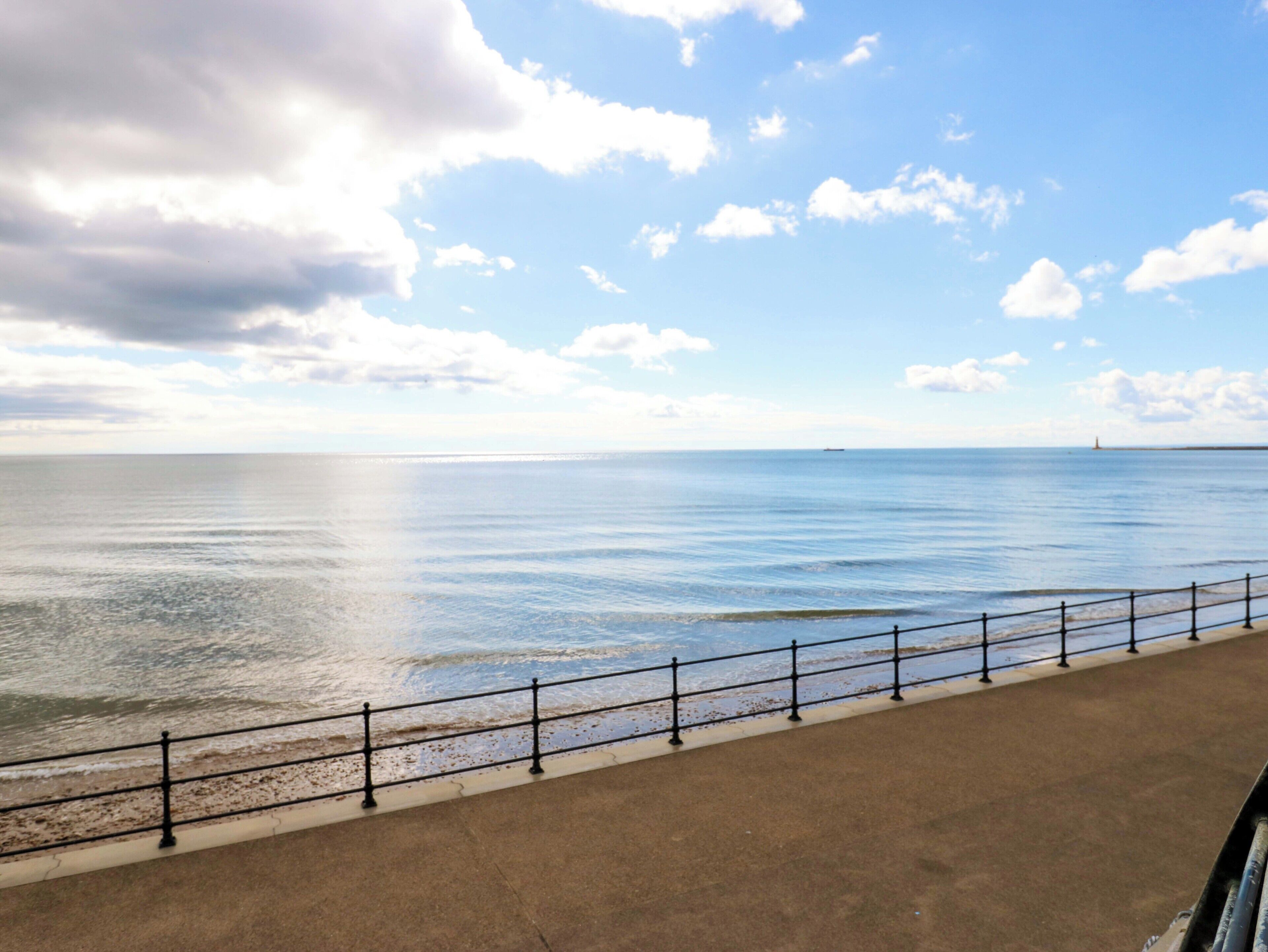 A coastal walkway with a metal railing, leading to a calm sea under a partly cloudy sky.