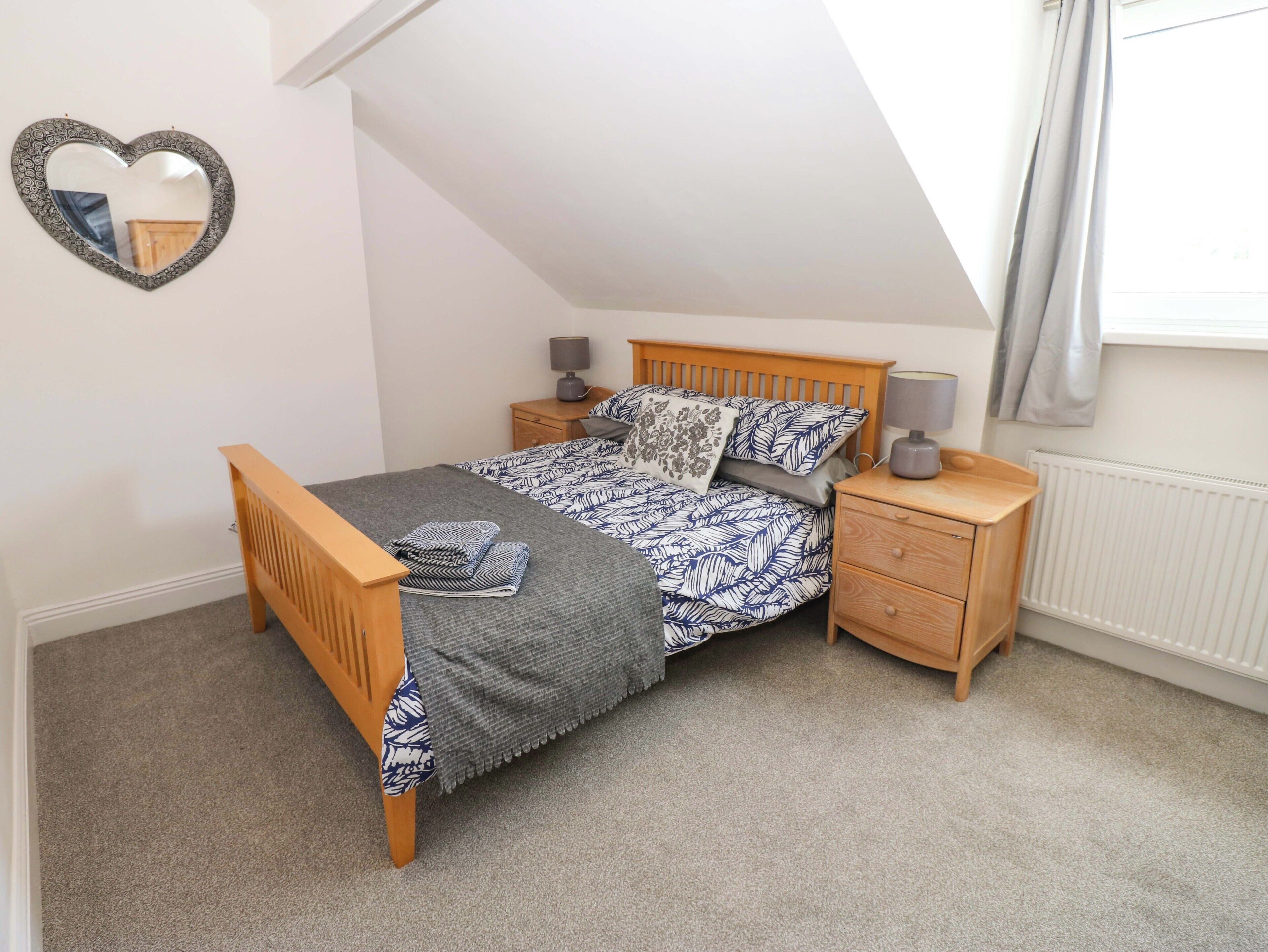 A bedroom with a sloped ceiling, a bed with patterned bedding, two bedside tables with lamps, a wooden nightstand, and a heart-shaped mirror on the wall.
