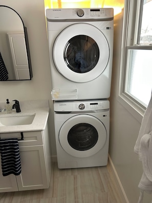 Un appareil compact combinant lave-linge et sèche-linge, doté d’un plan de travail et d’un évier blancs, d’un miroir et d’une fenêtre offrant une vue agréable.