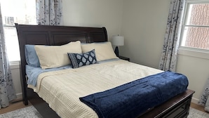 A bed with a quilted cover, a bedside table with a lamp, and a window with curtains. - Cozy, newly renovated home with a gorgeous fireplace and office space. (Midland)