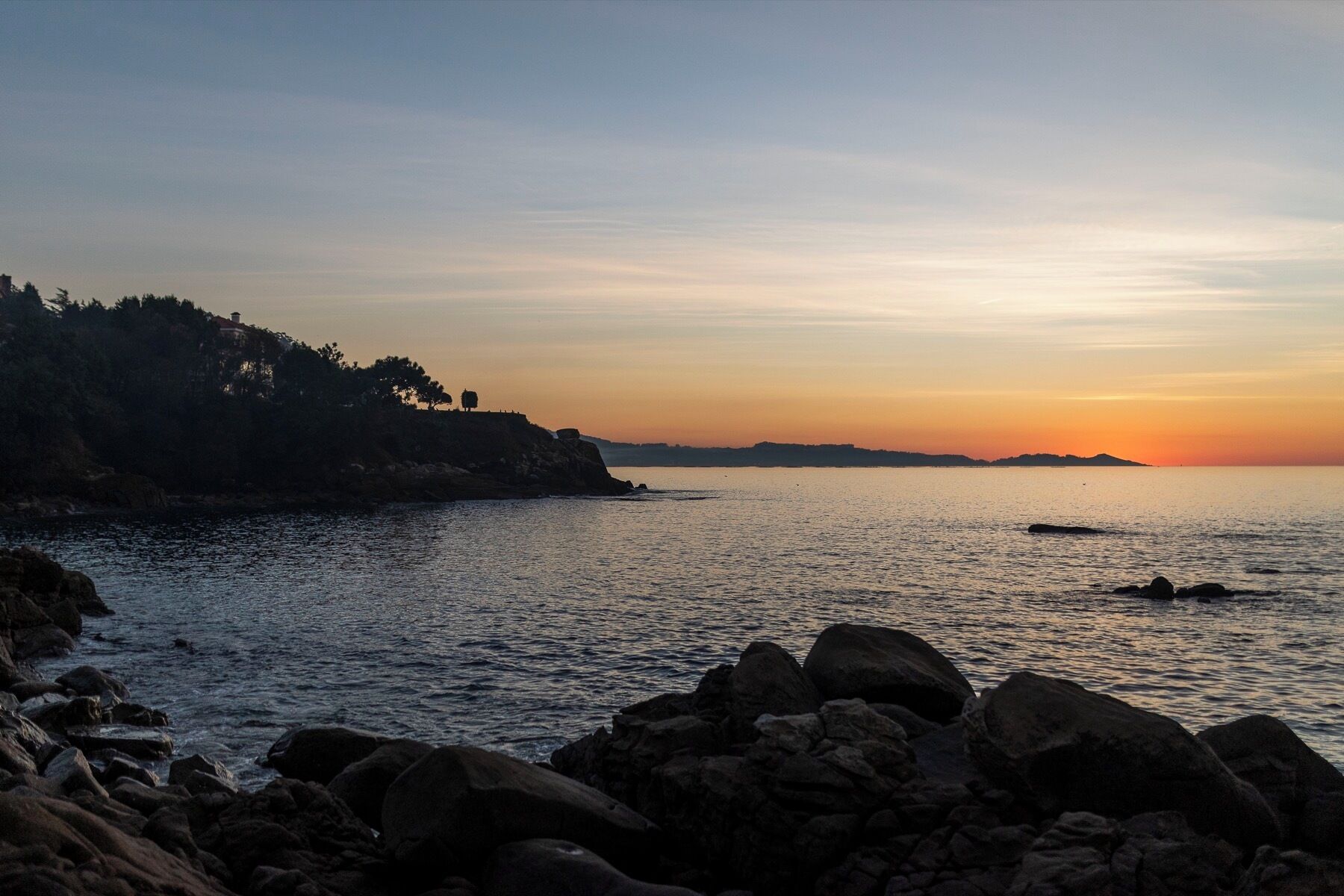 Paesaggio costiero con uno scoglio in primo piano, mare calmo e tramonto all'orizzonte.