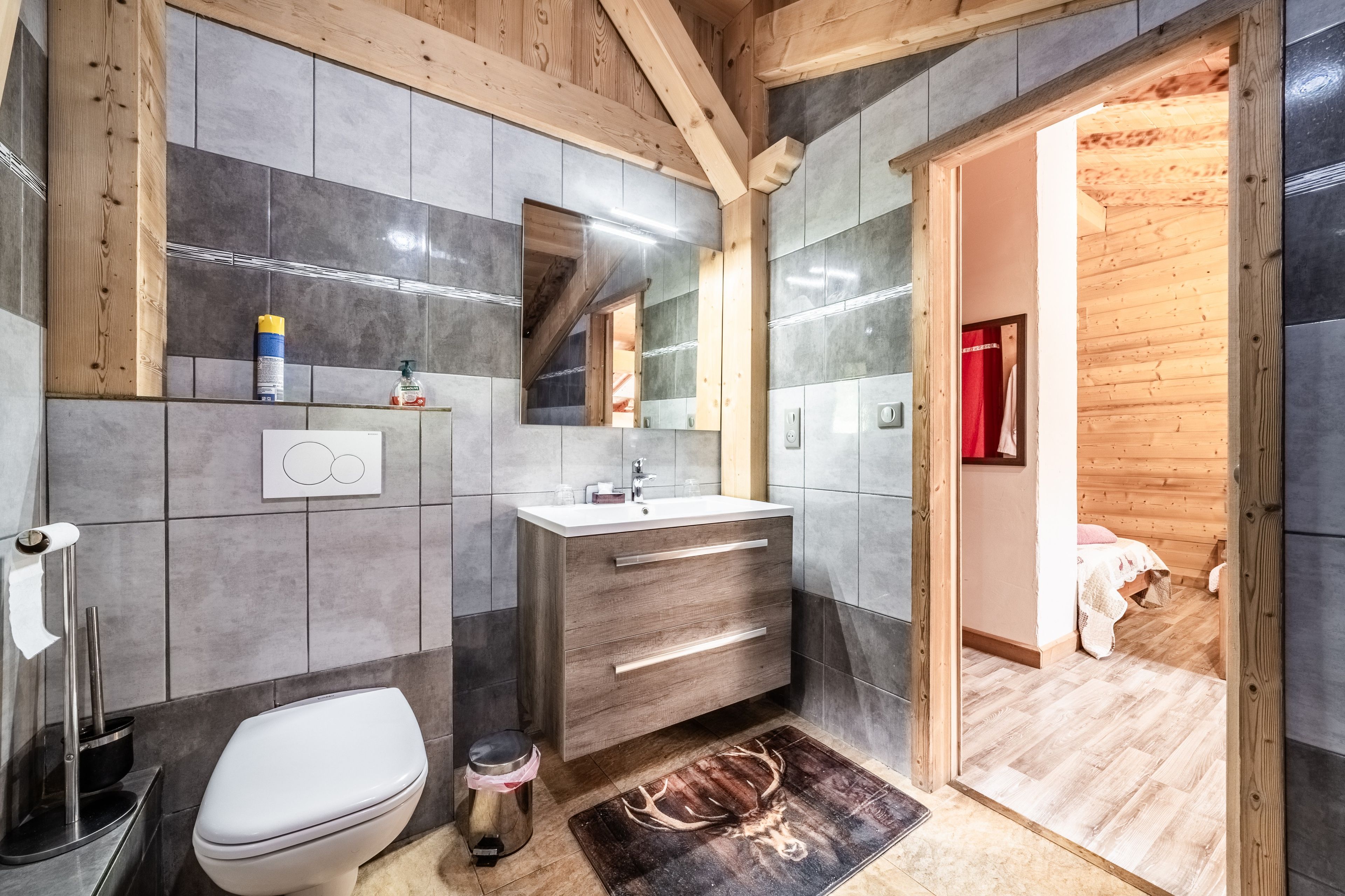 Et moderne badeværelse med træloft, grå fliser, et hvidt toilet, et spejl med vaskebord og et bruseområde.