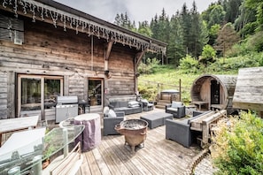 A wooden cabin with a deck, outdoor seating, a barbecue grill, and a wood stove.