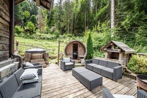 A wooden deck with wicker furniture, a hot tub, and a barrel-shaped structure in a forested area.