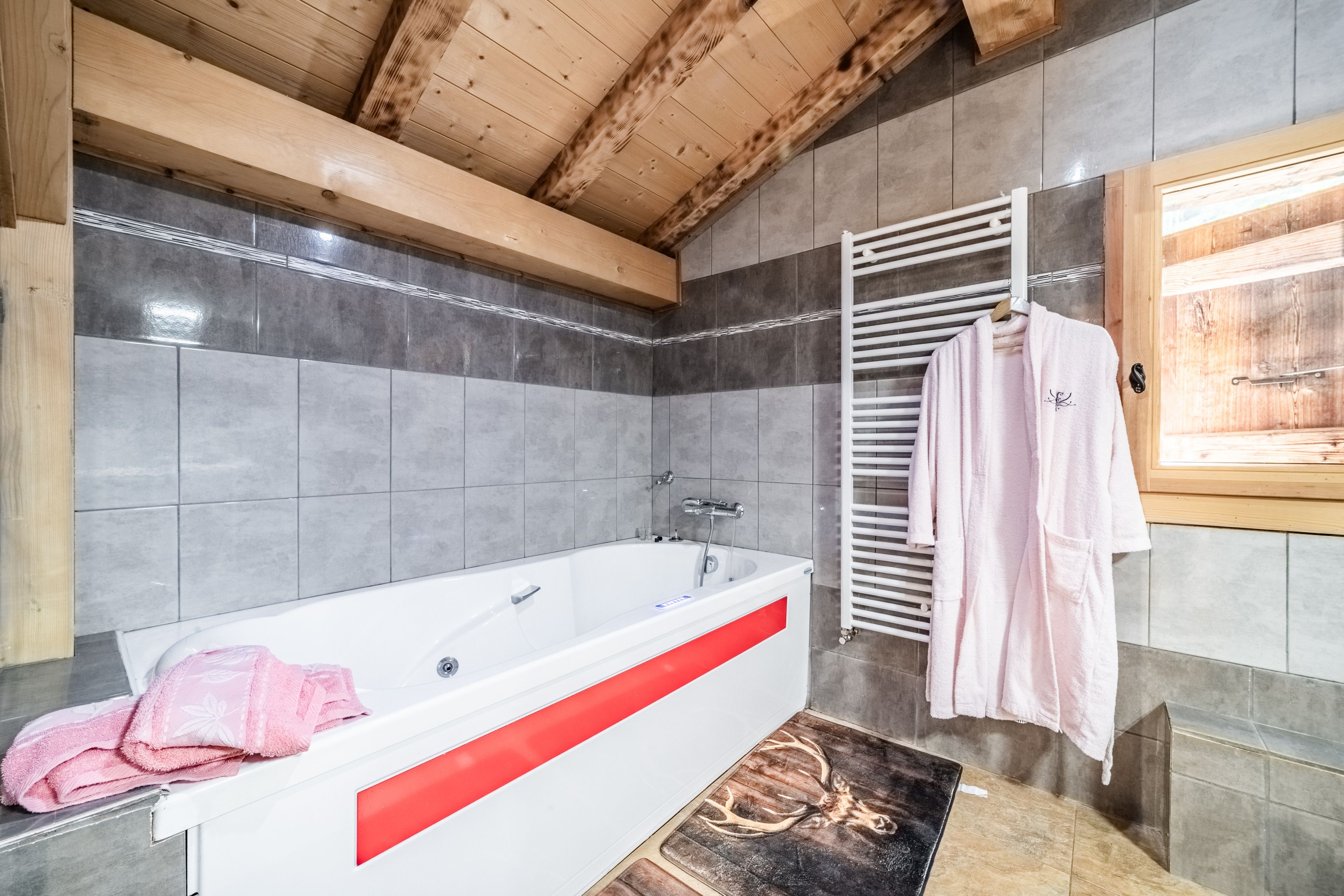 A modern bathroom with a large bathtub, grey tiles, wooden beams, and a towel rack with towels.