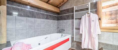 A modern bathroom with a large bathtub, grey tiles, wooden beams, and a towel rack with towels.